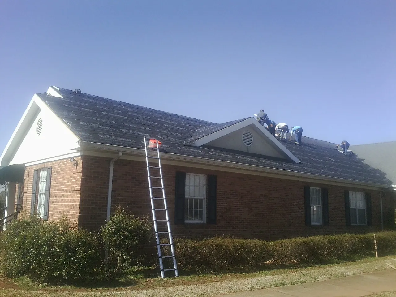 Crew tearing shingles off a brick ranch roof with a ladder set — Metal Roof Repair work by our crew in Mendon
