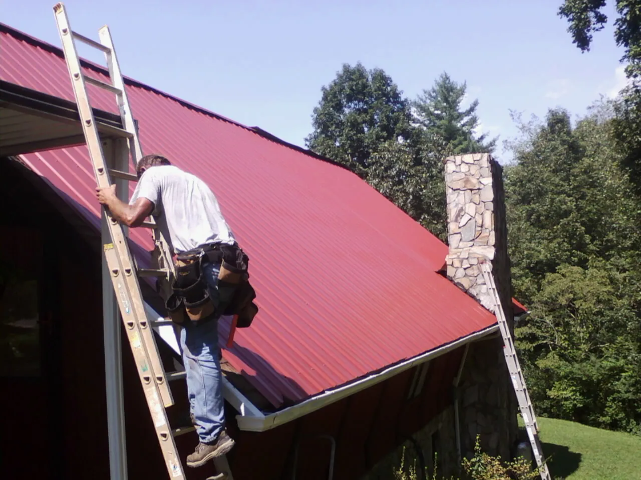 Roofer on a ladder beside a red metal roof and stone chimney — Soffit & Fascia Repair by Roofing Brothers in Mendon